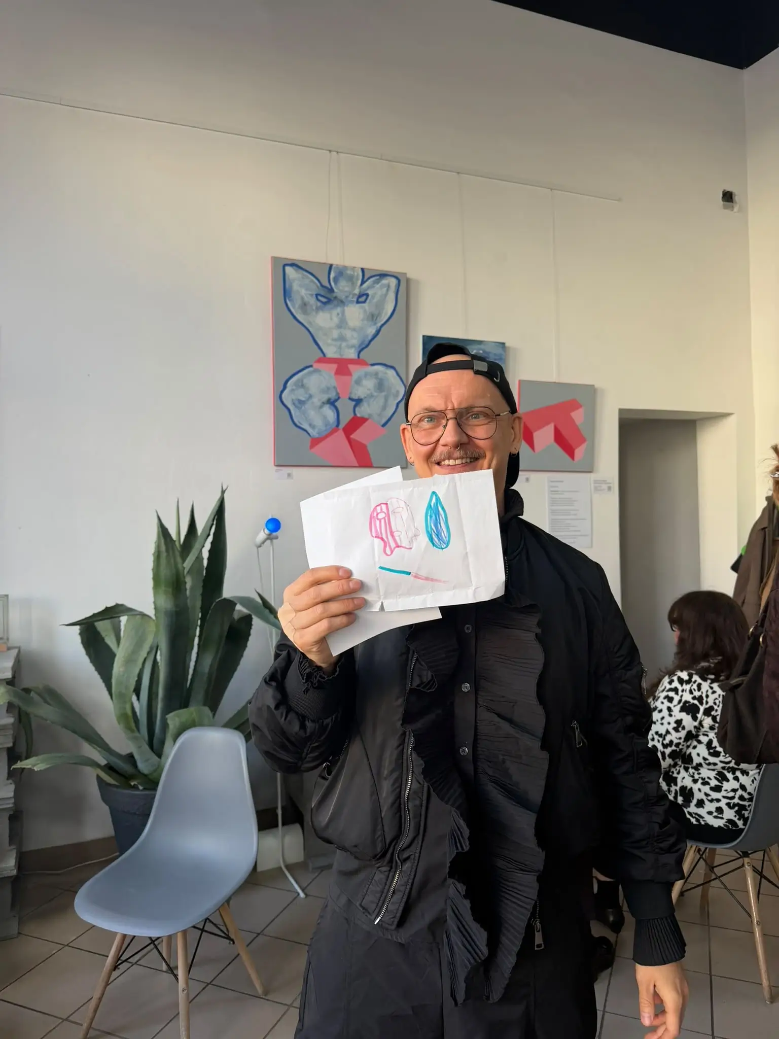 Mężczyzna w okularach pokazuje kolorowy dziecięcy rysunek na wystawie „Kolor Męskości” w Community Cup, w tle obrazy z serii na szarym tle