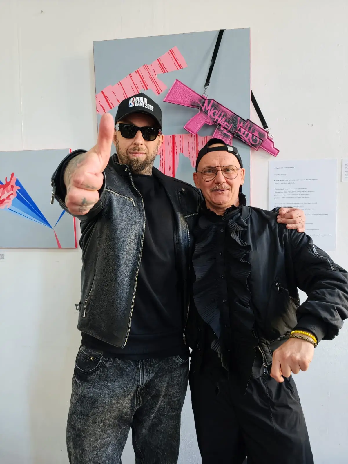 Dwóch mężczyzn pozuje przed obrazem z różowymi karabinami na wystawie „Kolor Męskości” w Community Cup, jeden pokazuje kciuk w górę